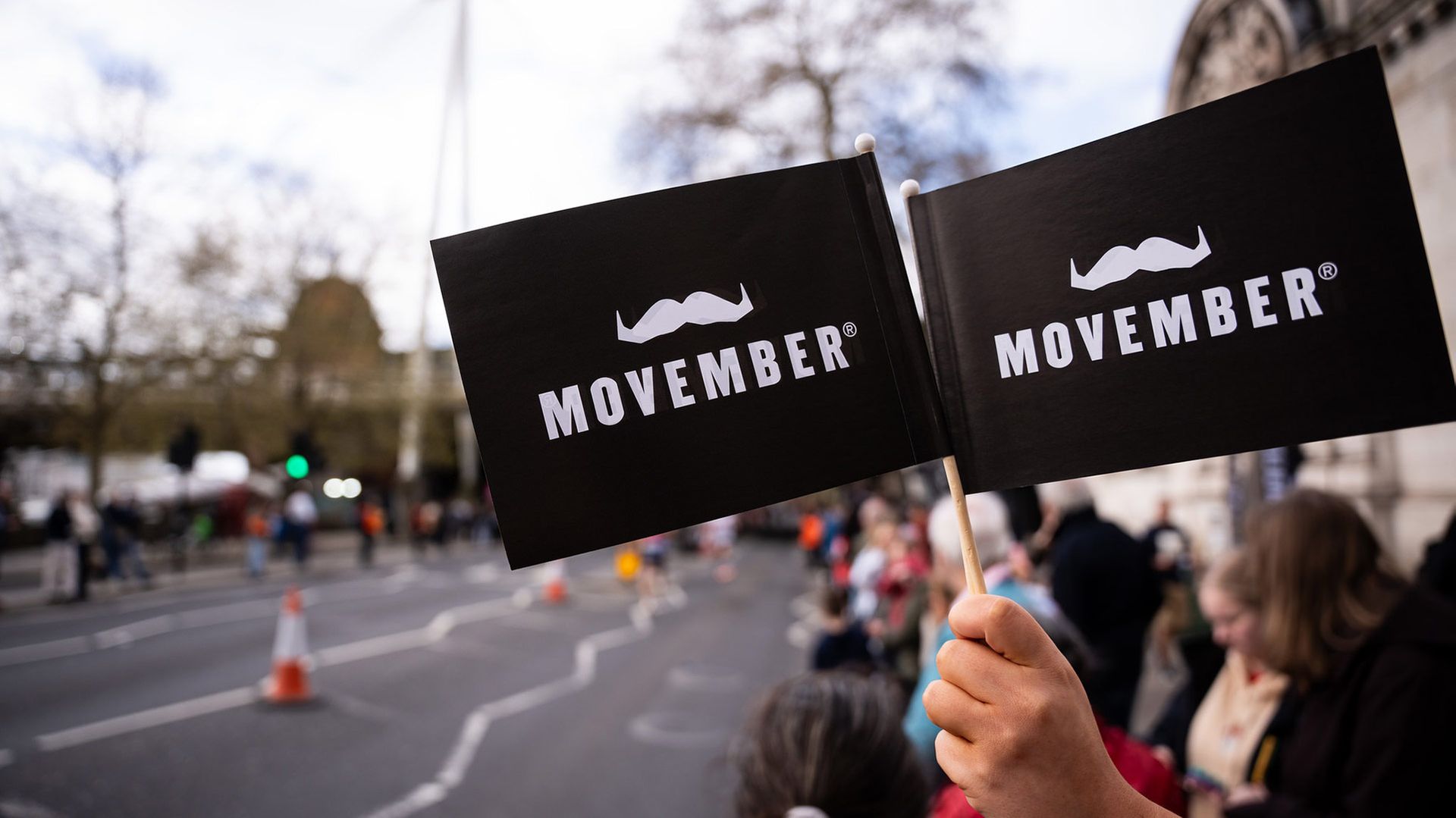 Photo of a hand holding two Movember-branded flags from the side-lines at a marathon.