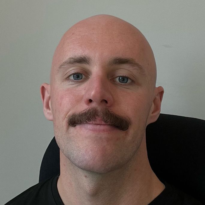 Portrait photo of a man with a striking moustache, smiling to camera.