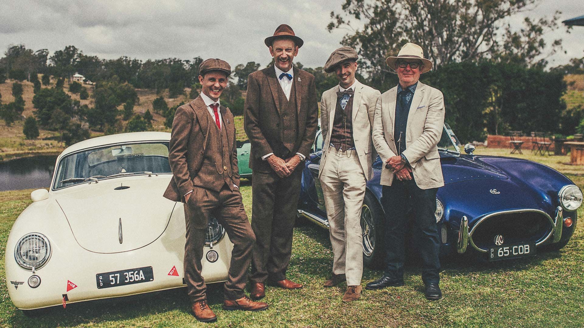 Photo of very well-dressed, dapper men, posing in front of classic cars.